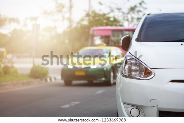 Focusing on the White car\
headlights on a street corner with sunlight flares and  small white\
car, In the background, the driver, Taxi, Bus and car. Closeup\
headlights of car.