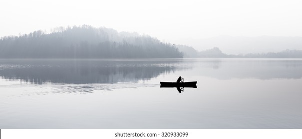 Fog over the lake. Silhouettes of trees. The man floats in a boat with a paddle. Black and white