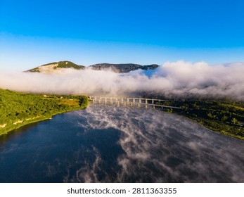 Fog rises over the river as sunlight breaks through in the morning. A bridge crosses the river while trees line the banks and mountains are visible in the distance.