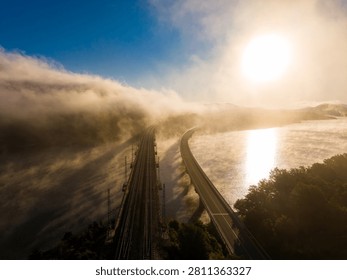 Fog surrounds the river and train tracks during sunrise in a rural area. The sun rises over the hills, creating a view of land and water.