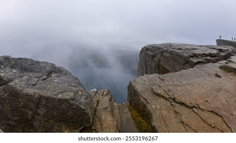 A foggy landscape featuring a rocky cliff overlooking a body of water, with mist obscuring the distant view. The scene is serene and atmospheric, with a few people standing on the cliff edge. - Powered by Shutterstock