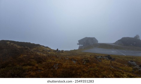 A foggy landscape with a modern building partially visible, surrounded by grassy hills and a parking area. The scene is enveloped in mist, creating a mysterious and serene atmosphere. - Powered by Shutterstock