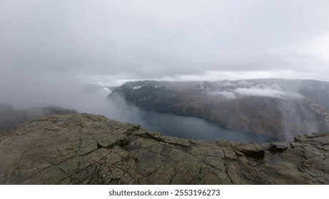 A foggy landscape with rocky cliffs overlooking a serene body of water, surrounded by mist and clouds. - Powered by Shutterstock