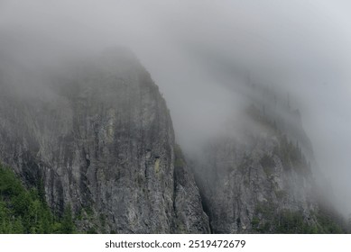 A foggy landscape shows towering, jagged cliffs partially obscured by thick mist. The cliff faces are rugged with steep rocky surfaces, and sparse evergreen trees cling to the sides. The scene feels m - Powered by Shutterstock