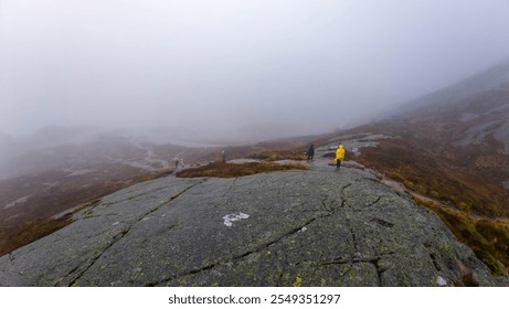 A foggy mountain landscape with hikers on a rocky path, one wearing a yellow jacket. - Powered by Shutterstock