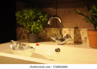 A Foil Toy Boat With A Red Flag Floats In A Sink With A Tap And Home Flowers