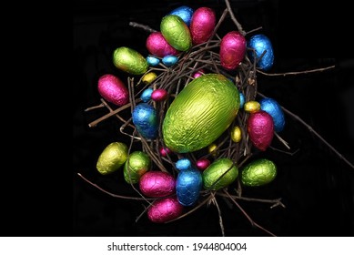 Foil wrapped multi coloured easter eggs in pink, green, blue and yelow in a natural nest made of wood, sticks and twigs, against a black background. 