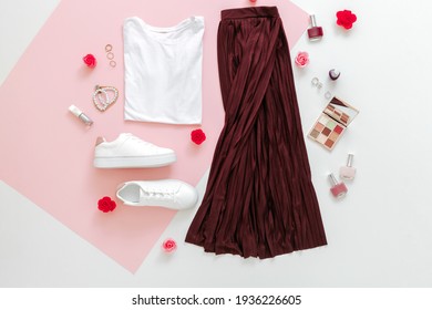 Folded Clothes For Women Fashion Urban Basic Outfit With Accessories Flowers Make Up Cosmetics On Pink Background.Female Spring Look Summer Outfit Skirt Shoes Sneakers Basic Tshirt Bag. Top View.