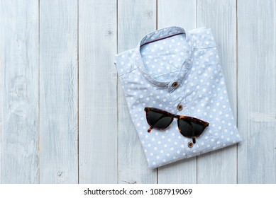 Folded Light Blue Polkadot Linen Shirt With Sunglasses On White Wooden Table Background.