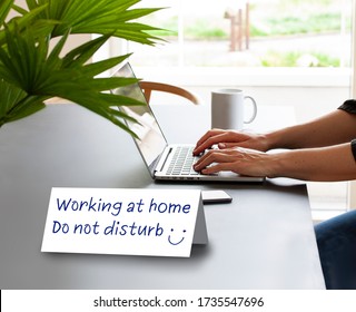 Folded Paper Card Standing On A Table Saying Working At Home - Do Not Disturb. Hands Of A Person Working On A Laptop Computer With Coffee Mug And A Mobile Phone. Working Or Studying Concept. 