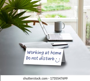 Folded Paper Card Standing On A Table Saying Working At Home - Do Not Disturb. Laptop Computer On A Table With Coffee Mug, Notepad, Mobile Phone And Pencil. Working Or Studying At Home Concept.