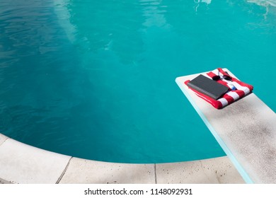 Folded Red And White Striped Beach Towel Folded On A Diving Board With A Book, Sunscreen, And Aviator Sunglasses On Top. Swimming Pool In The Backyard With Folded Towel And Book, No People.