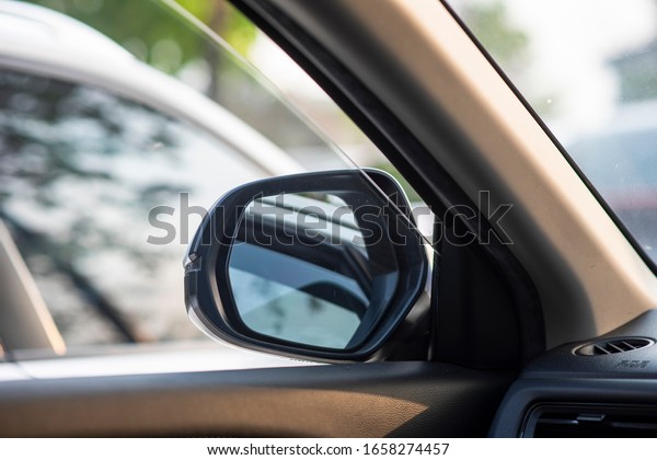 Folded wing mirror of a compact modern car, with a\
side window glass drop down for a ventilation, parked in a parking\
lot during the morning time with a nice sun rise shining. Seeing\
from a back seat.