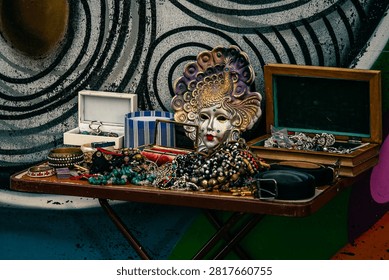 A folding table is covered with an eclectic display of vintage jewelry and accessories for sale, including layered bracelets, beaded and stone necklaces, a black belt with buckle, and several open jewelry boxes. At the center, a decorative Venetian-style mask with gold and purple feathered details and colorful faux gemstones stands out among the pile, with a small mirror propped up in front of it. The scene is set against a painted wall featuring a bold concentric circle pattern in black, white, and turquoise, adding an artistic urban backdrop. Warm, directional light highlights the metallic and glass textures of the jewelry, creating a rich, textured still life. This image works well for flea market, vintage shopping, antique jewelry, and eclectic decor themes.