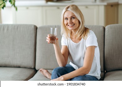 Follow healthy lifestyle. Senior caucasian attractive blonde dressed in a casual wear sits on the couch in a living room, holding a glass of pure water in a hand looks at the camera and smiling - Powered by Shutterstock