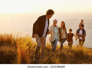 Follow the leader. A multi-generational family walking up a grassy hill together at sunset with the ocean in the background.