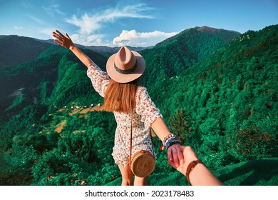 Follow me concept and traveling together. Girl traveler wearing hat, round straw bag and short jumpsuit holds the boyfriend's hand and leading to green big mountains vacation 