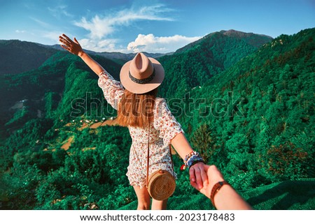 Follow me concept and traveling together. Girl traveler wearing hat, round straw bag and short jumpsuit holds the boyfriend's hand and leading to green big mountains vacation 