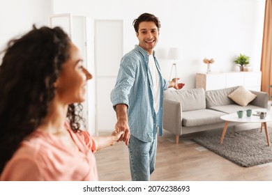 Follow Me. Portrait of excited guy inviting lady to enter home, welcoming to living room, happy smiling man holding woman's hand showing new modern apartment and living room, selective focus