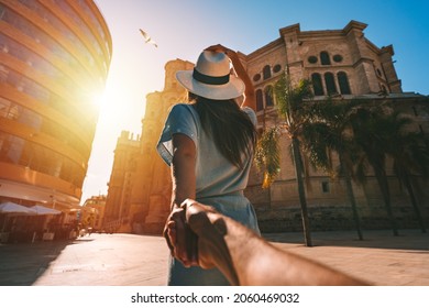 Follow me - POV. Young tourist woman in white sun hat holding her boyfriend by hand and walking in Malaga city at sunset. Couple on summer holiday vacation in Spain. Traveling together. - Shutterstock ID 2060469032