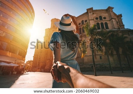 Follow me - POV. Young tourist woman in white sun hat holding her boyfriend by hand and walking in Malaga city at sunset. Couple on summer holiday vacation in Spain. Traveling together.