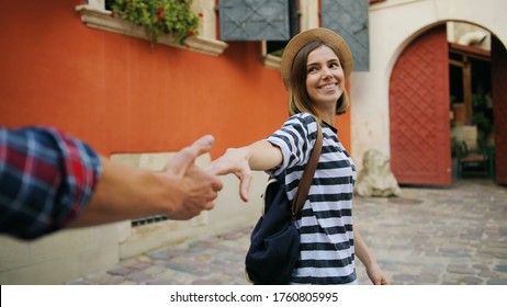 Follow Me Shot Of Attractive Young Woman In A Straw Hat Holding Man's Hand And Showing Him The Landmarks Of The City. Caucasian Couple Walking Happily Down The Medieval Street.