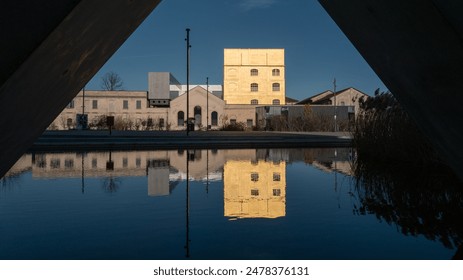 Fondazione Prada, a striking architectural marvel in Milan, stands reflected in a tranquil pool, its tower and buildings bathed in a warm, golden glow, creating a serene and contemplative atmosphere. - Powered by Shutterstock