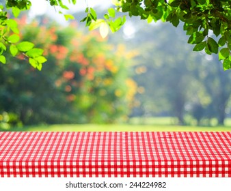 Food Background, Empty Picnic Table And Red Tablecloth Over Blur Green Tree Park With Bokeh In Spring Summer Nature Background, Product Display Montage Mock Up, Template, Banner, Backdrop, Wallpaper