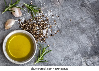 Food Background With Olive Oil, Peppercorns, Sea Salt, Rosemary, And Garlic Cloves, Over Dark Slate Background.  Overhead View.