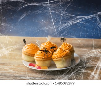 Food, Baking And Holidays Concept - Cupcakes Or Muffins With Halloween Party Decorations, Jelly Worm Candy And Spiderweb On Wooden Table Over Starry Night Sky Background