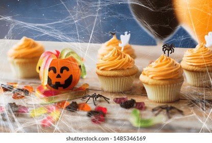 Food, Baking And Holidays Concept - Cupcakes Or Muffins With Halloween Party Decorations, Jelly Candies And Spiderweb On Wooden Table Over Starry Night Sky Background