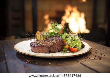 Food - Beef dinner - Delicious grilled stake and potatoes served on a wooden table, fireplace on background. Big steak meat dish on a main course plate
