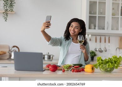 Food Blog, Enjoy Cooking, Take Photos On Modern Device, Video Call. Smiling Young African American Lady Drinking Wine, Making Selfie On Phone In Kitchen Interior, With Laptop And Vegetables On Table