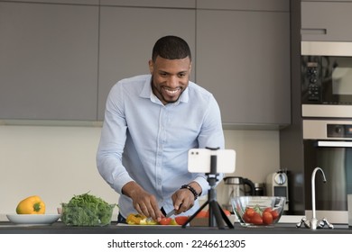 Food blog, on-line recipes. African vlogger record cooking class on smartphone camera, watch tutorial prepare vegetarian dish, cuts fresh paprika and tomato stand in kitchen looks at cellphone screen - Powered by Shutterstock