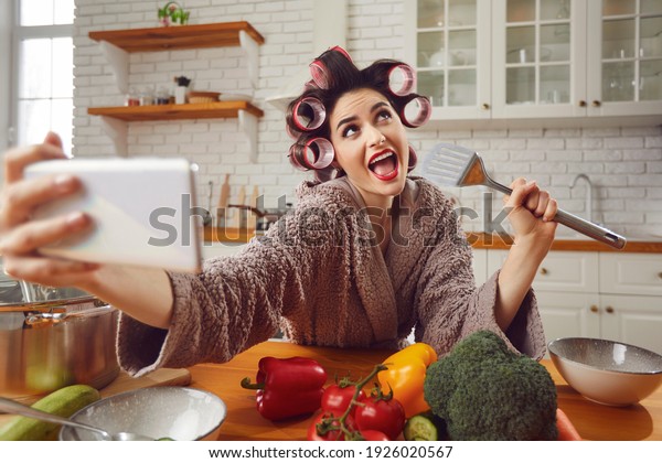 Food blogger, singer or social media addict having\
fun while cooking meal in kitchen. Young woman in hair curlers and\
housecoat singing in pretend spatula mic and taking funny selfie on\
mobile phone