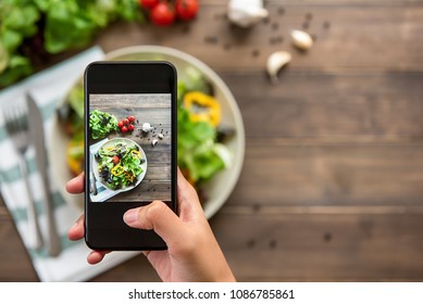 Food blogger using smartphone taking photo of beautiful mix fresh green salad on wood table to share on social media