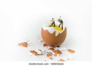 Food, Business Concept. Two Miniature Figures Worker Stand And Woking On Brown Egg To Break Eggshell And Digging To Yolk - On White Background