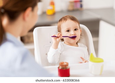 food, child, feeding and people concept - little baby girl with spoon sitting in highchair and eating puree from jar and mother at home kitchen - Powered by Shutterstock