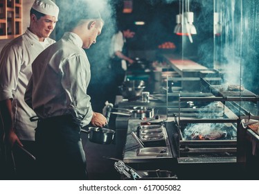 Food concept. Chef in white uniform monitors the degree of roasting and greases meat with oil in saucepan in interior of modern restaurant kitchen. Preparing traditional beef steak on barbecue oven.