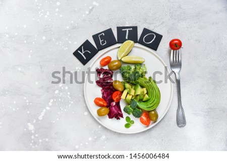 food concept for keto diet: avocado and broccoli, tomatoes and olives, leaves radicchio and lime. on white craft plate. on a black plate letters KETO. healthy lunch. ketogenic diet. food flat lay 
