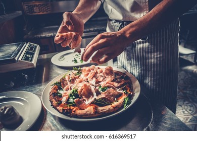 Food concept. Preparing traditional italian pizza. Chef in uniform decorate ready dish with green rucola herbs, ham, salami and prosciutto in interior of modern restaurant kitchen. Ready to eat. - Powered by Shutterstock