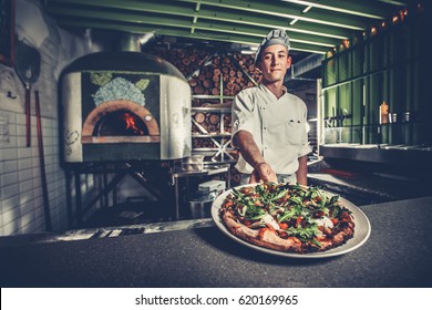 Food concept. Preparing traditional italian pizza. Young smiling chef in white uniform and gray hat show ready dish with green rucola herbs in interior of modern restaurant kitchen. Ready to eat.