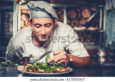 Food concept. Preparing traditional italian pizza. Young smiling chef in white uniform and gray hat decorate ready dish with green rucola herbs in interior of modern restaurant kitchen. Ready to eat.