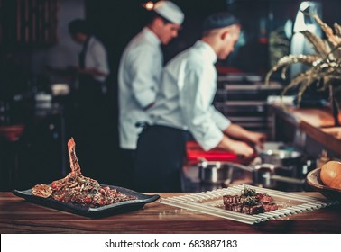 Food Concept. Ready Grilled Pork Ribs Dish And Beef Steak With Herbs. Ready To Serve. Ready To Eat. Two Chefs Working In The Background Interior Of Modern Professional Restaurant Kitchen