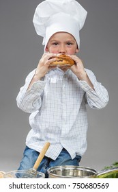 Food Concepts. Portrait Of Happy Caucasian Little Boy Eating Fresh Bun With Cinnamon. Posing In Cooking  Hat Agaunst Gray Background. Vertical Image Composition