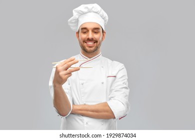 Food Cooking, Asian Cuisine And People Concept - Happy Smiling Male Chef In Toque Holding Something With Chopsticks Over Grey Background (focus On Chopsticks)