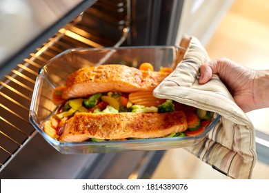 Food Cooking, Culinary And People Concept - Young Woman With Potholder Taking Baking Dish With Salmon Fish And Vegetables Out Of Oven At Home Kitchen