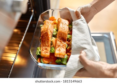 Food Cooking, Culinary And People Concept - Young Woman With Towel Taking Baking Dish With Salmon Fish And Vegetables Out Of Oven At Home Kitchen