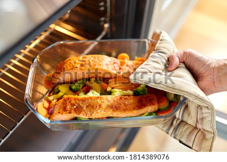 food cooking, culinary and people concept - young woman with potholder taking baking dish with salmon fish and vegetables out of oven at home kitchen