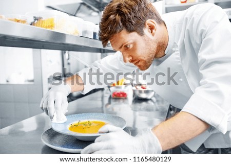 food cooking, profession and people concept - happy male chef cook serving and cleaning plate of soup at restaurant kitchen